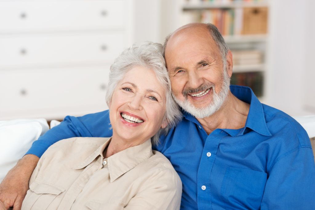 Romantic senior couple sitting close together on a sofa in the house smiling happily and you can't tell which one has dental implants. 