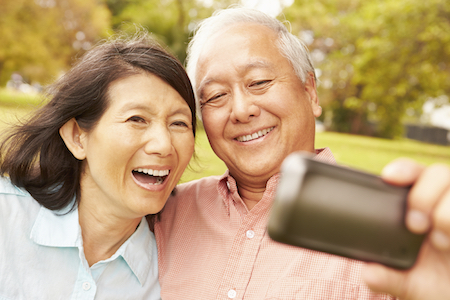 Senior Asian Couple Taking Selfie In Park Together with Perfect Smiles from Dental Impants.
